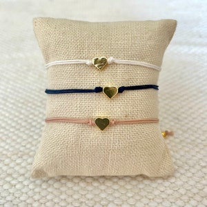Puede incluir: Tres pulseras con dije de corazón en un expositor de tela blanca. Las pulseras están hechas con hilo blanco, azul marino y rosa. Cada pulsera tiene un dije de corazón dorado.