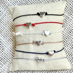 Könnte beinhalten: Ein Set aus sechs Armbändern mit silbernen Herzanhängern. Die Armbänder bestehen aus verschiedenen farbigen Schnüren, darunter Schwarz, Rot, Weiß, Beige, Dunkelblau und Grau.
