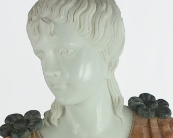 May include: A white marble bust of a woman with a detailed hairstyle, wearing a carved, peach-colored bodice with green floral accents. The bust is mounted on a black base.