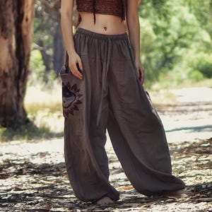 Puede incluir: Un pantalón holgado de lino gris con un bolsillo decorativo que presenta un diseño de sol y ojo. El pantalón tiene una cintura con cordón y lo lleva puesto una persona de pie en un entorno boscoso.