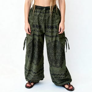Könnte beinhalten: Olivgrüne Hose mit schwarzem geometrischen Muster. Die Hose hat einen Kordelzug in der Taille, Taschen und Bindebänder an den Knöcheln. Die locker sitzende Hose ist aus einem weichen, fließenden Stoff gefertigt.