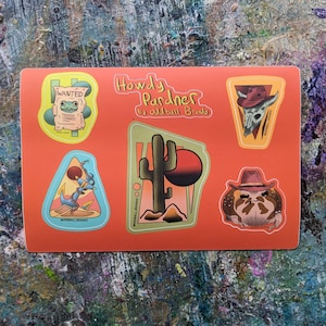 May include: An orange sticker sheet featuring five distinct designs: a frog on a wanted poster, a skull wearing a cowboy hat, a cactus with a sunset, a lassoed anchor, and a frog wearing a cowboy hat. The text Howdy Pardner by Oddball Brando is centered.