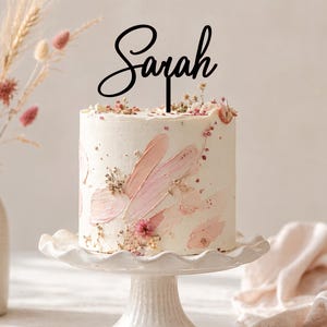 May include: A black cake topper featuring the name Sarah in a cursive script font, positioned atop a cylindrical white cake decorated with pink brushstroke frosting patterns and small dried flowers, resting on a white pedestal cake stand.