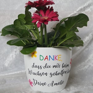 Könnte beinhalten: Weißer Keramik-Blumentopf mit einer bunten "DANKE"-Botschaft und dem Text "dass du mir beim Wachsen geholfen hast" und "Deine Amelie". Der Topf ist mit kleinen gelben, rosa und blauen Blumen verziert. Der Topf enthält eine rosa Gerbera-Pflanze.