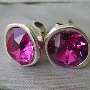 Può includere: Un paio di orecchini a bottone argentati. Ogni orecchino presenta una grande gemma sfaccettata color magenta incastonata in una lunetta. Le gemme sono tagliate per riflettere la luce. Gli orecchini sono esposti su uno sfondo neutro. Gioielli.