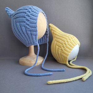 Może przedstawiać: Dwa dziane czepki dla niemowląt z troczkami. Jeden czepek jest niebieski, a drugi żółty.