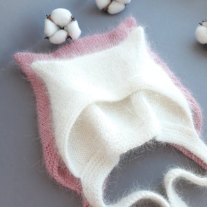 Op de afbeelding: Een witte gebreide baby muts met een roze rand en een pluche textuur. De muts heeft een strik aan de achterkant.