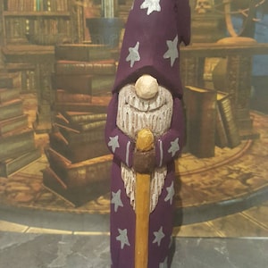 May include: A purple wooden gnome figurine wearing a pointed hat with silver stars. The gnome is holding a wooden staff and has a white beard.