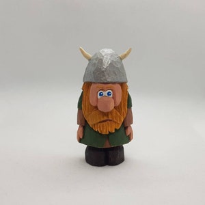 Pode incluir: Uma figura de madeira de um viking com uma túnica verde, uma barba castanha e um capacete prateado com chifres. A figura tem olhos azuis e um nariz grande.
