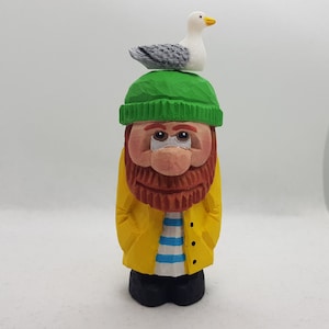 May include: A wooden figurine of a fisherman wearing a yellow raincoat, a green hat, and a white and blue striped shirt. A white seagull is perched on top of the hat.