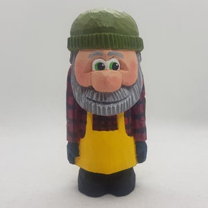 Può includere: Una statuetta in legno intagliata a mano che rappresenta una persona con un cappello verde, una camicia a quadri rossi e neri e una salopette gialla. La statuetta ha una barba grigia e un'espressione amichevole.