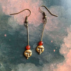 May include: A pair of gold dangle earrings with a small red bead and a gold moon charm with a face on each earring.