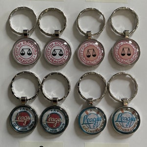 Peut inclure: Quatre ensembles de porte-clés avec des motifs différents. Les deux premiers ensembles présentent un charme rond avec le texte "City of Rockford Peaches" et un motif de balance de la justice. Les deux derniers ensembles présentent un charme rond avec le texte "A League of Their Own" et un motif de baseball.