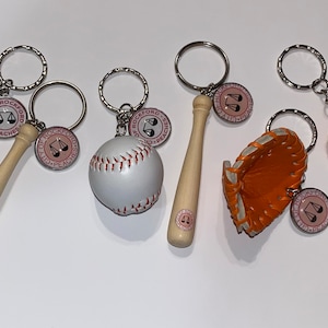 Op de afbeelding: Vijf sleutelhangers met baseball-thema's, waaronder een honkbal, een knuppel en een vanghandschoen. Elke hanger is bevestigd aan een zilveren sleutelring met een ronde hanger waarop "City of Rockford Peaches" staat.