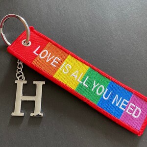Op de afbeelding: Een rode, gele, groene, blauwe en roze gestreepte sleutelhanger met de tekst "Love is all you need" in wit. De sleutelhanger heeft een zilveren hanger in de vorm van de letter H.