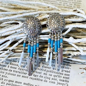 May include: A pair of silver-toned dangle earrings featuring an oval concho top with a central floral motif. Each earring has four hanging strands composed of small silver beads, bright turquoise cylindrical beads, and flat silver metal tips.