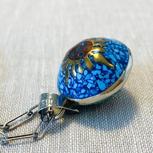 May include: A pendant featuring a dome-shaped inlay of crushed blue turquoise stone with a central gold-toned sun motif. The piece is set in a silver-toned metal frame and attached to a silver-toned metal chain link necklace with a stamped bail.