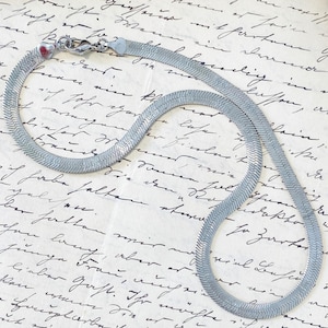 May include: A silver-toned herringbone chain necklace featuring a flat, flexible link design with a polished finish. The necklace is secured with a lobster claw clasp and includes small heart-shaped end caps at the connection points of the chain.