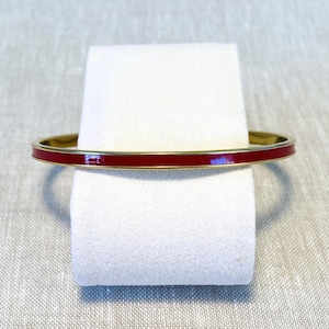 May include: A thin, circular bangle bracelet featuring a vibrant red enamel inlay centred between two polished gold-tone metal edges. The design is sleek and uniform, showcasing a smooth, glossy finish that reflects light along the entire circumference.