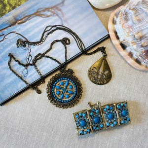 Può includere: Un set di gioielli color bronzo composto da una lunga collana a catena con un grande ciondolo circolare con cabochon blu, un ciondolo separato a forma di goccia con dettagli intagliati e un bracciale a cerniera decorato con cabochon blu.