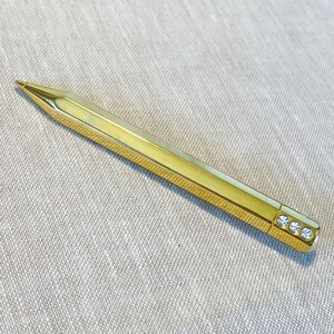 May include: A gold-toned metal pen with a hexagonal barrel and a pointed tip. The top end of the pen features three small, clear, circular crystals embedded in a row, providing a decorative accent to the sleek, polished metallic surface.