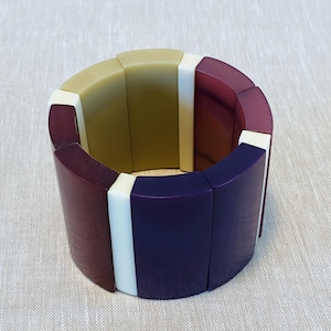 Op de afbeelding: Een brede armband bestaande uit afwisselend gebogen segmenten in olijfgroene, diepbordeauxrode en donkerpaarse tinten, gescheiden door dunne, verticale witte rechthoekige tussenstukjes, wat een gesegmenteerd, veelkleurig cirkelvormig ontwerp vormt.