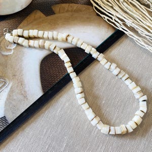 May include: A necklace composed of numerous small, off-white, cube-shaped beads with subtle tan variations. The beads are strung together in a single strand, featuring a metallic silver-toned clasp at one end to secure the jewelry piece.