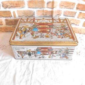 Könnte beinhalten: Rechteckige Blechdose mit goldenem Rand und detaillierter Illustration einer Winterszene mit Menschen und einer Bäckerei. Der Deckel hat Text, und die Dose steht vor einer Backsteinmauer.