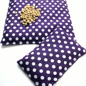May include: Two purple fabric heating pads with white polka dots. The larger pad has a pile of beige beans on top.
