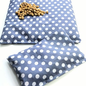 May include: Two gray and white polka dot fabric heating pads. The larger pad is lying flat and the smaller pad is folded in half. A pile of light brown beans is on top of the larger pad.