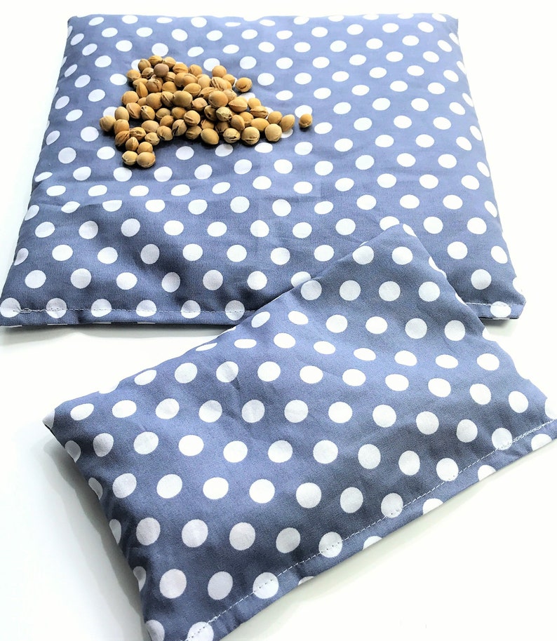 May include: Two gray and white polka dot fabric heating pads. The larger pad is lying flat and the smaller pad is folded in half. A pile of light brown beans is on top of the larger pad.