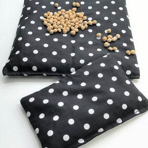 May include: Two black fabric hot packs with white polka dots. The larger hot pack has a scattering of beige beans on top.