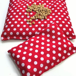 May include: Two red and white polka dot fabric heat packs. The larger pack is lying flat and the smaller pack is folded in half on top of the larger pack.