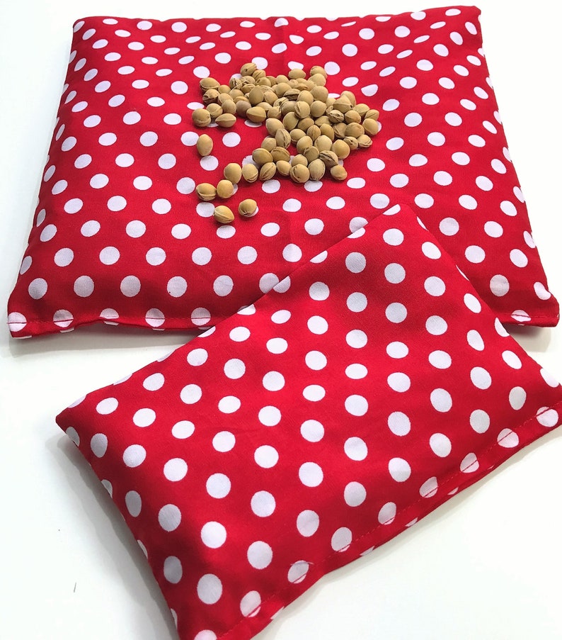 May include: Two red and white polka dot fabric heat packs. The larger pack is lying flat and the smaller pack is folded in half on top of the larger pack.
