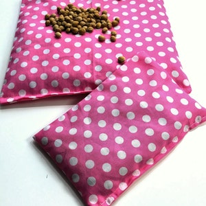 May include: Two pink polka dot fabric heat packs. The larger pack has a scattering of brown seeds on top.