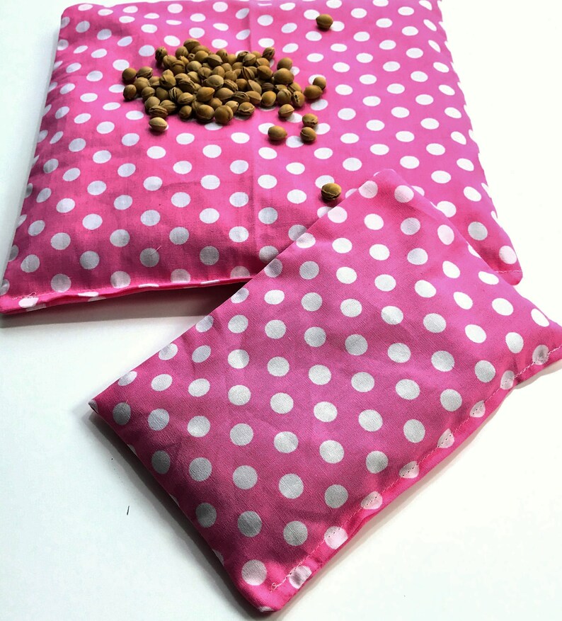May include: Two pink polka dot fabric heat packs. The larger pack has a scattering of brown seeds on top.