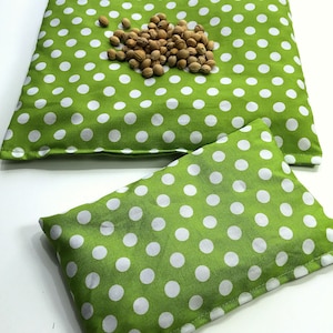 May include: Two green and white polka dot fabric heat packs. The larger pack is lying flat and the smaller pack is on top of it.