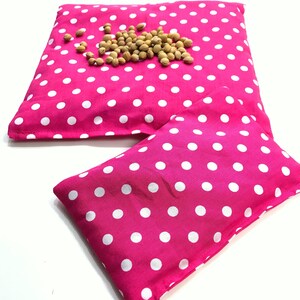 May include: Two pink polka dot fabric heat packs. The larger pack has a pile of brown flax seeds on top.