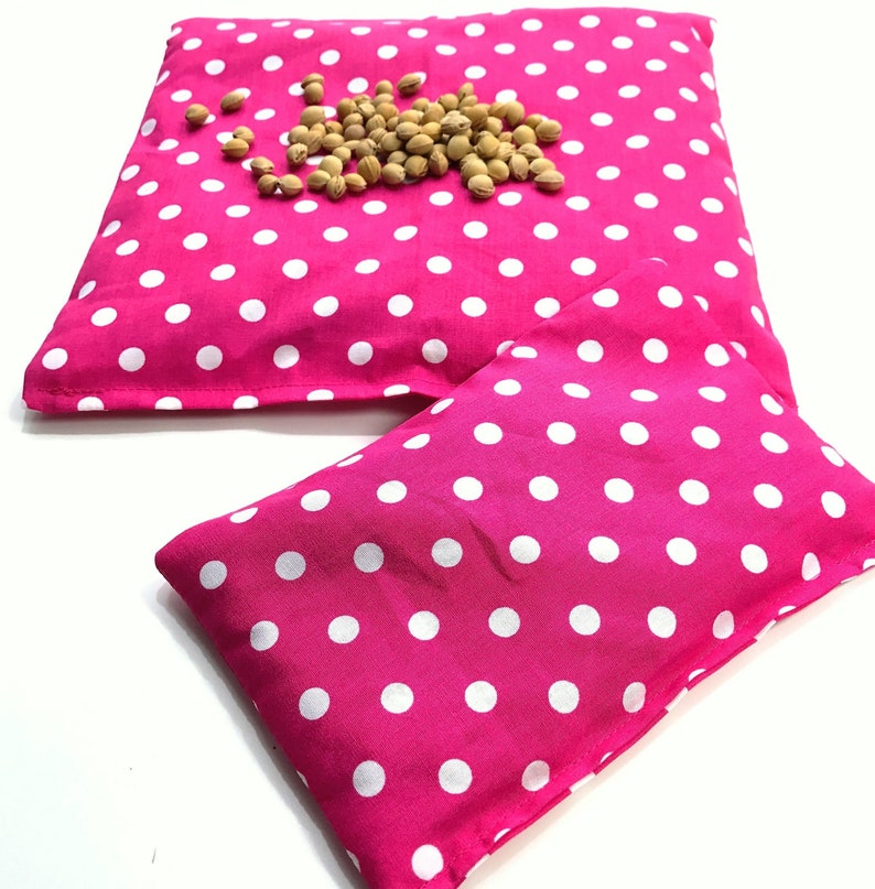 May include: Two pink polka dot fabric heat packs. The larger pack has a pile of brown flax seeds on top.