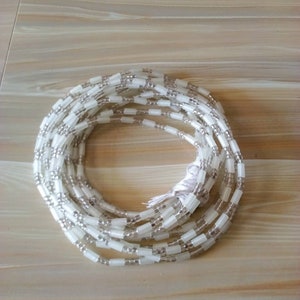 Op de afbeelding: Een witte kralen tailleketting met zilveren kralen. De ketting is gemaakt van kleine, ronde kralen en grotere, rechthoekige kralen.