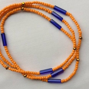 Op de afbeelding: Een tailleketting gemaakt van oranje en blauwe kralen. De ketting is gemaakt van kleine, ronde oranje kralen en grotere, rechthoekige blauwe kralen. Er zijn ook kleine gouden kralen verspreid over de ketting.