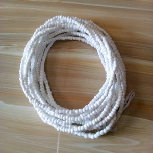 Op de afbeelding: Een witte kralen tailleketting, ook wel bekend als een taillekraal of buikketting, is gemaakt van kleine, ronde kralen die aan elkaar geregen zijn op een koord. De ketting is opgerold op een houten oppervlak.