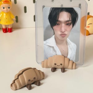 May include: A clear plastic card holder with a photo of a person with dark hair and a white shirt. Two brown croissant-shaped card holders with smiling faces are in the foreground. A yellow duck figurine and a cartoon character are in the background.