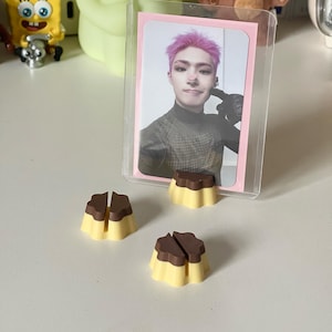 May include: A photograph of a person with pink hair in a clear plastic card holder, with a pink border. Three yellow and brown pudding-shaped card holders are in front of the photo. Other figurines are in the background.