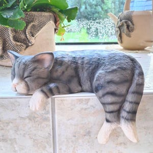 Könnte beinhalten: Grau-weiße Katzenfigur in Schlafposition. Die Katze hat die Augen geschlossen und ruht auf einer gefliesten Oberfläche. Die Figur hat ein gestreiftes Muster und weiße Pfoten. Eine Topfpflanze und eine dekorative Teekanne befinden sich im Hintergrund.