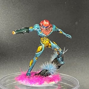 May include: A hand-painted miniature figurine of a blue and yellow space suit character with a red helmet. The character is standing on a pink and blue fuzzy base.
