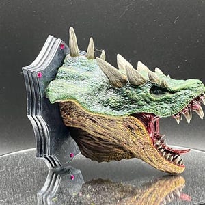 May include: A green and brown dragon head sculpture with a fierce expression, mounted on a black and silver wall plaque with pink gems. The dragon's mouth is open, revealing sharp teeth and a red tongue.