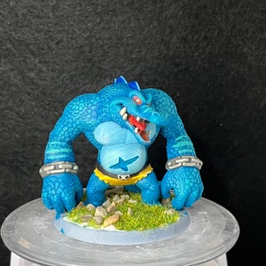 May include: A blue and green painted miniature figurine of a monster with a crocodile head, wearing a yellow loincloth and a silver chain bracelet. The figurine is standing on a base with green grass and rocks.