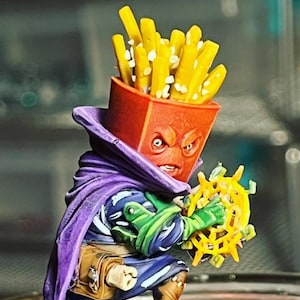 May include: A small, painted figurine of a character wearing a purple cape and holding a yellow and orange object. The character has a red, angry-looking face with a yellow and white french fry container on top of its head.