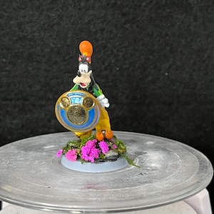 May include: A hand-painted miniature figurine of a cartoon dog wearing a green and yellow outfit, holding a gold shield with a blue circle and three gold circles. The figurine is standing on a base with pink flowers and green foliage.